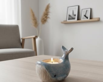 May include: A blue and white ceramic whale-shaped candle holder with a lit tea light inside. The whale has a textured body and a small eye detail. The candle holder is on a light-colored wooden table.