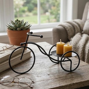 May include: Black metal bicycle-shaped candle holder with two yellow candles. The decorative piece sits on a wooden table, with a succulent in a terracotta pot and eyeglasses in the background. The candle holder has a circular tray.
