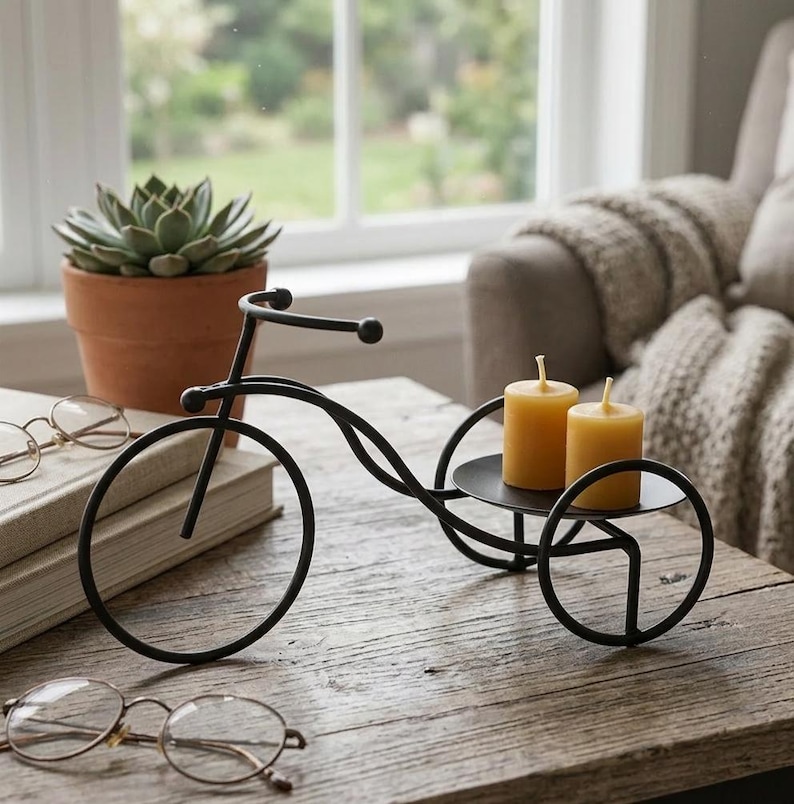 May include: Black metal bicycle-shaped candle holder with two yellow candles. The decorative piece sits on a wooden table, with a succulent in a terracotta pot and eyeglasses in the background. The candle holder has a circular tray.