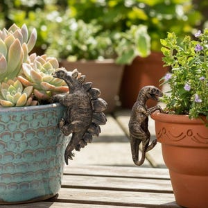 May include: Bronze dinosaur ornaments clinging to plant pots. A Stegosaurus decorates a turquoise pot, and a Velociraptor clings to a terracotta pot. Other potted plants and greenery are visible in the background.