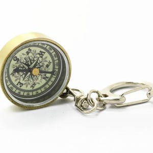 K&ouml;nnte beinhalten: Ein Kompass-Schl&uuml;sselanh&auml;nger im Vintage-Stil mit einem messingfarbenen Geh&auml;use und einem silberfarbenen Verschluss. Das Kompasszifferblatt hat detaillierte Richtungsmarkierungen und eine Nadel. Ideal f&uuml;r Navigation und Reisen.