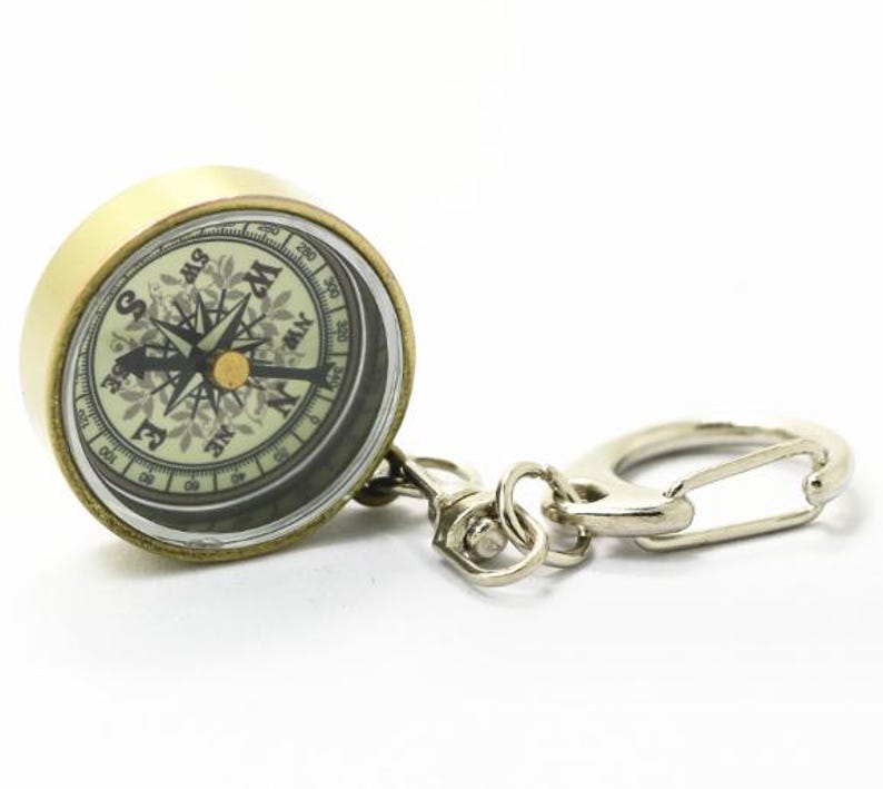 K&ouml;nnte beinhalten: Ein Kompass-Schl&uuml;sselanh&auml;nger im Vintage-Stil mit einem messingfarbenen Geh&auml;use und einem silberfarbenen Verschluss. Das Kompasszifferblatt hat detaillierte Richtungsmarkierungen und eine Nadel. Ideal f&uuml;r Navigation und Reisen.
