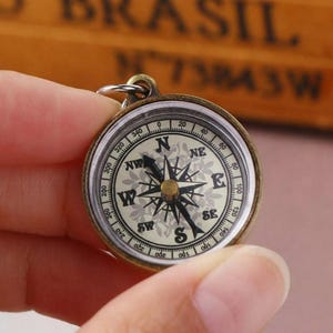 K&ouml;nnte beinhalten: Ein Kompass im Vintage-Stil mit einem messingfarbenen Geh&auml;use. Das Kompasszifferblatt zeigt ein detailliertes Design mit Richtungsmarkierungen (N, S, O, W) und Gradanzeigen. Der Kompass wird von einer Hand gehalten.