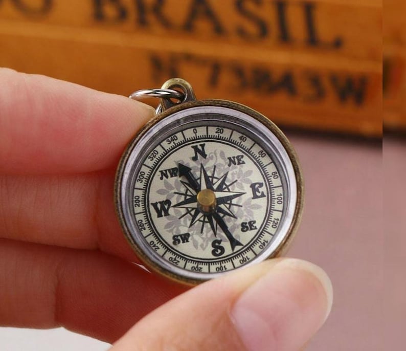 K&ouml;nnte beinhalten: Ein Kompass im Vintage-Stil mit einem messingfarbenen Geh&auml;use. Das Kompasszifferblatt zeigt ein detailliertes Design mit Richtungsmarkierungen (N, S, O, W) und Gradanzeigen. Der Kompass wird von einer Hand gehalten.