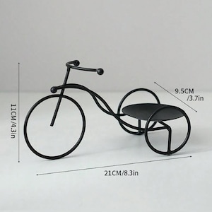 May include: Black metal tricycle-shaped decorative piece. The front wheel is 4.3 inches (11 cm) in diameter, and the entire piece is 8.3 inches (21 cm) long. The top platform is 3.7 inches (9.5 cm) wide. The design is minimalist and modern.