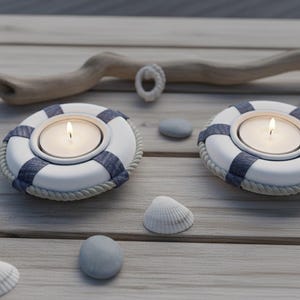 May include: Two white and navy blue life preserver candle holders with lit candles. The candle holders have a rope trim and are surrounded by seashells and stones on a wooden surface. A driftwood piece is in the background.