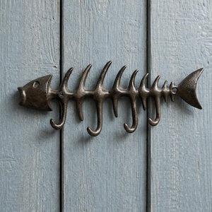 May include: A decorative wall hook in the shape of a fish skeleton, made from cast iron. The hook has a dark, metallic finish and is mounted on a light blue wooden surface. The design includes a fish head, tail, and several curved hooks.