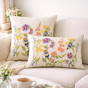Può includere: Due cuscini decorativi con ricami floreali e di uccelli. I cuscini presentano uno sfondo color crema con fiori colorati viola, gialli e arancioni e piccoli uccelli. I cuscini hanno un bordo verde.