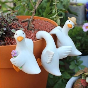 Può includere: Statuine di anatra in ceramica bianca con becco e zampe arancioni, progettate per essere appese al bordo di un vaso di terracotta. Un'anatra tiene in mano un fiore viola e un'altra indossa un piccolo cappello di paglia. Decorazioni per il giardino.