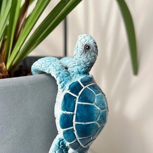 Puede incluir: Una figura de tortuga de cerámica azul decora una maceta gris. La tortuga tiene un caparazón detallado y está pintada en tonos de azul y blanco. Se ven hojas verdes de plantas en el fondo.