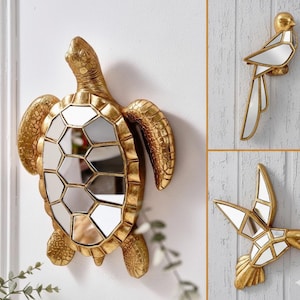May include: Gold-coloured wall mirrors shaped like a turtle, a bird, and a hummingbird. The turtle mirror has a geometric pattern, while the bird and hummingbird mirrors have mirrored sections. The mirrors are mounted on a white wall.