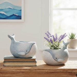 May include: Two whale-shaped ceramic planters in light blue with white accents. One planter is empty, and the other contains purple and white flowers. The planters sit on a wooden table with books and a painting in the background.