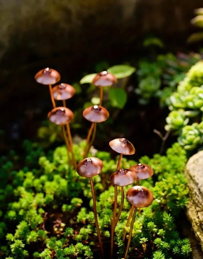 May include: A cluster of miniature copper-colored mushroom sculptures with rounded caps and slender stems, emerging from a bed of vibrant green moss. The mushrooms have a reflective surface, catching the light. The scene evokes a natural, whimsical aesthetic.