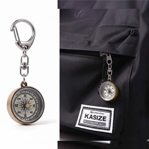 K&ouml;nnte beinhalten: Ein Kompass-Schl&uuml;sselanh&auml;nger im Vintage-Stil mit einem silbernen Karabiner und einer Kette. Der Kompass hat ein goldfarbenes Geh&auml;use und ein detailliertes Zifferblatt mit Richtungsmarkierungen. Der Kompass ist an einem schwarzen Rucksack mit einem "KASIZE"-Etikett befestigt.