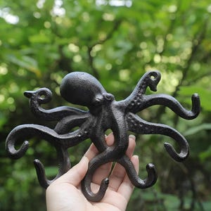 May include: A black cast iron octopus wall hook with multiple curved arms and hooks. The octopus has a textured surface and is held in a person's hand against a blurred green background.