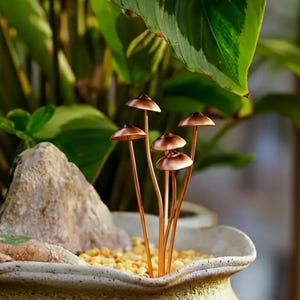 May include: A decorative arrangement featuring several copper-colored mushroom sculptures. The mushrooms have small, rounded caps and slender stems, placed in a ceramic pot with small yellow stones. Lush green foliage provides a natural backdrop.