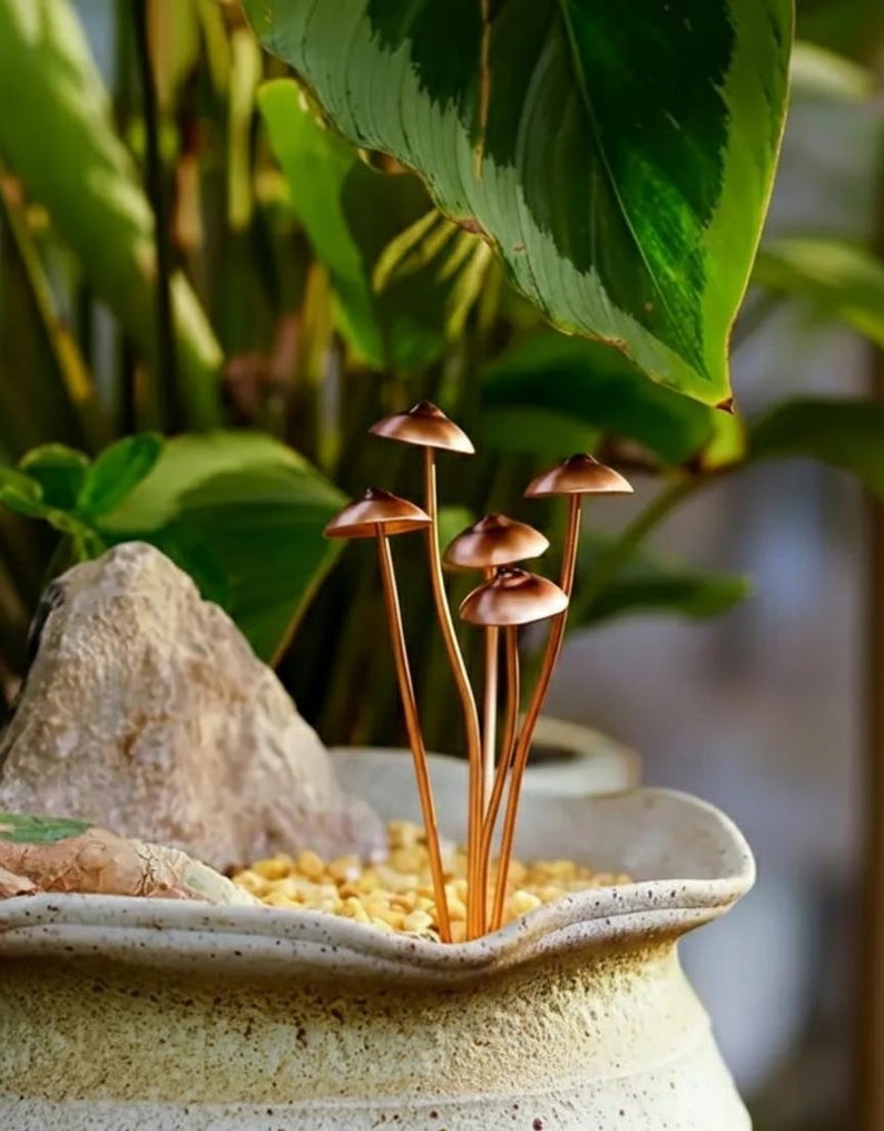 May include: A decorative arrangement featuring several copper-colored mushroom sculptures. The mushrooms have small, rounded caps and slender stems, placed in a ceramic pot with small yellow stones. Lush green foliage provides a natural backdrop.