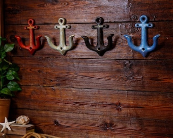 Puede incluir: Cuatro ganchos de pared con forma de ancla en rojo, blanco roto, negro y azul. Los ganchos están montados en una pared rústica de madera oscura. Decoración de temática náutica.