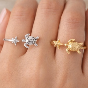 May include: Two adjustable rings, one silver and one gold, each with a starfish and turtle design. The rings are shown on a hand, highlighting the jewellery's details. The turtle has a textured shell, and the rings are adjustable.