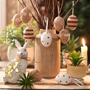 May include: Easter decorations including a white rabbit ornament, a beige vase with branches holding hanging Easter eggs, and a small potted succulent. The eggs are decorated with patterns and ribbons. The scene is set on a table.