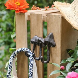 May include: A set of three cast iron mushroom-shaped hooks mounted on a wooden slat. A blue and white patterned bag hangs from one hook. A straw hat rests on the wooden structure, with an orange flower and greenery in the background.