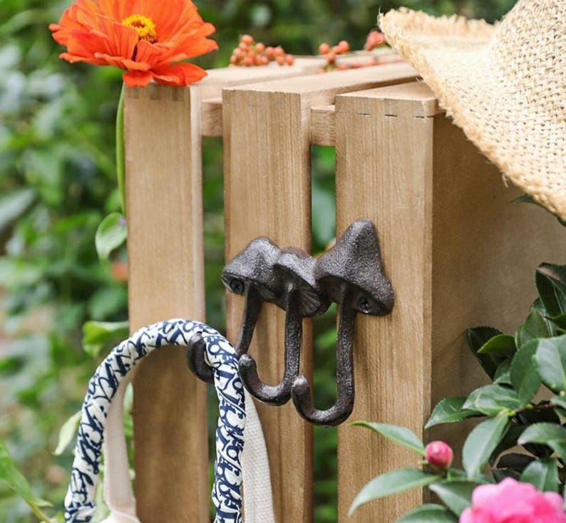 May include: A set of three cast iron mushroom-shaped hooks mounted on a wooden slat. A blue and white patterned bag hangs from one hook. A straw hat rests on the wooden structure, with an orange flower and greenery in the background.