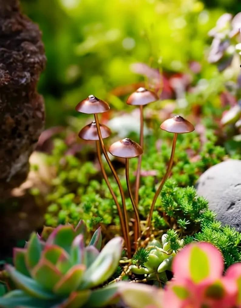 May include: A close-up shot of several copper-colored mushroom sculptures with rounded caps and slender stems. The mushrooms are set amidst a vibrant garden of green succulents and other plants, creating a whimsical, natural scene.