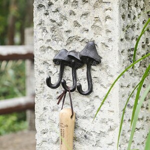 May include: A black cast iron wall hook with three mushroom-shaped heads and curved hooks. The hooks are mounted on a textured, light gray stone wall. A wooden item with text hangs from one of the hooks.