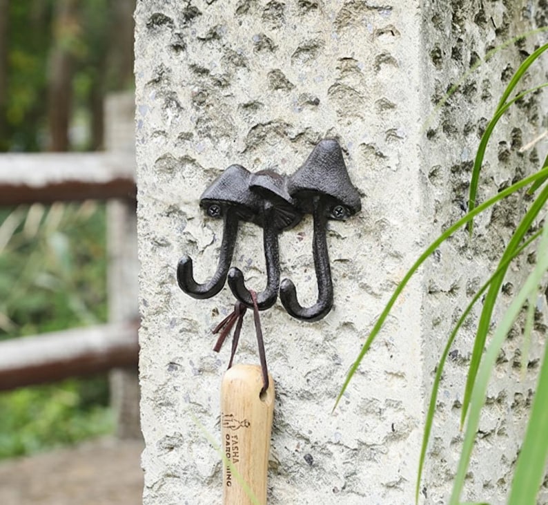 May include: A black cast iron wall hook with three mushroom-shaped heads and curved hooks. The hooks are mounted on a textured, light gray stone wall. A wooden item with text hangs from one of the hooks.