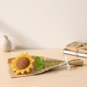 May include: A handmade crochet sunflower with yellow petals and a brown centre, presented as a bouquet. The flower is wrapped in newspaper and tied with a white ribbon, creating a charming, rustic aesthetic.