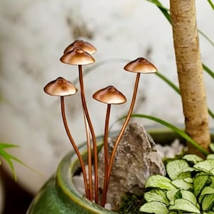 May include: Copper-toned mushroom sculptures with rounded caps and thin stems are arranged in a green ceramic pot. The mushrooms are placed among greenery and a small rock, creating a miniature garden scene. The pot is placed near a tree trunk.