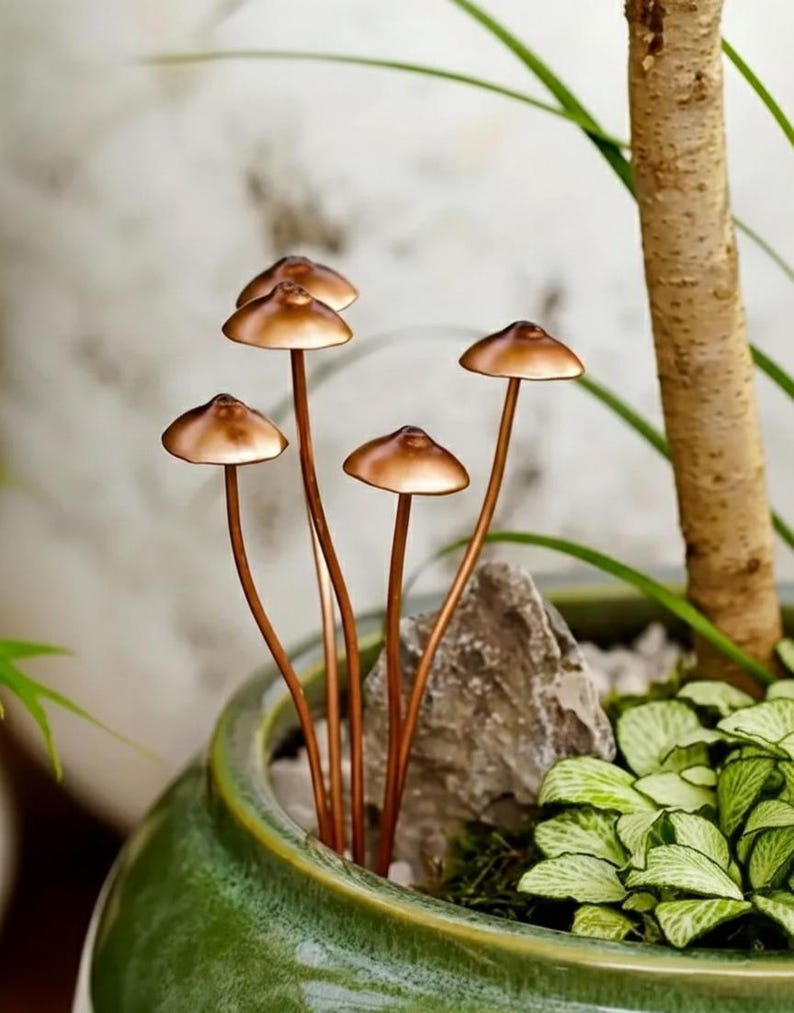 May include: Copper-colored mushroom sculptures with rounded caps and slender stems are arranged in a green ceramic pot. The mushrooms are placed among greenery and a small rock, creating a miniature garden scene. The pot is placed near a tree trunk.