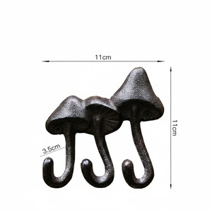 May include: A cast iron wall hook with three mushroom-shaped heads and curved hooks. The dark brown hook measures 11 cm in height and width, with the hooks extending 3.5 cm. The decorative hook is designed for hanging items.