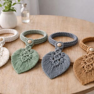 May include: Four macramé leaf-shaped towel rings in cream, green, grey, and tan, each with a wooden button detail. The rings are displayed on a round wooden table, showcasing a bohemian home decor style.