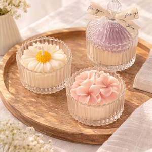 May include: Three decorative candles in glass containers on a wooden tray. One candle is shaped like a daisy, another like a flower, and the third is in a jar with a lid and a ribbon that says "for you".
