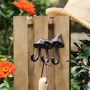 May include: Three cast iron mushroom-shaped hooks mounted on a wooden fence. The hooks are black and have a rustic finish. A wooden handled item hangs from the hooks. The background includes orange flowers, green foliage, and a straw hat.