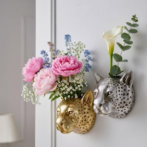 May include: Two wall-mounted vases shaped like leopard heads. One is gold, the other silver. The gold vase holds pink peonies and other flowers. The silver vase holds a white calla lily and greenery. Decorative home decor.