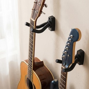 May include: Two black wall-mounted guitar hangers. One holds a brown acoustic guitar, the other a blue electric guitar. The guitars are mounted on a beige wall near a window with a white curtain.