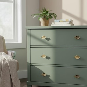 May include: A sage green chest of drawers with three drawers, adorned with gold leaf-shaped handles. A small potted plant and books are on top. The room has a window and neutral-toned walls.