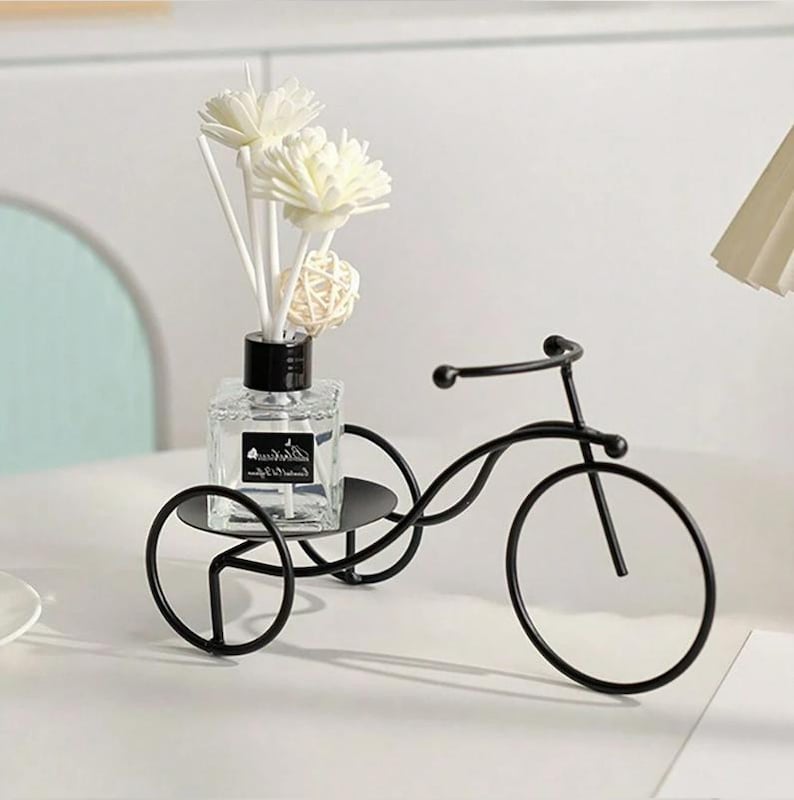 May include: A decorative black metal tricycle holds a clear glass bottle with a black cap and white reeds. The bottle contains a liquid and is topped with white flower-shaped decorations. A small, woven ball is also in the bottle.