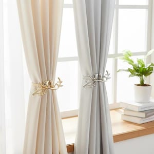 May include: Two sets of curtains, one cream and one grey, are held back by decorative metal curtain tiebacks. The tiebacks feature a bird perched on a branch design. A houseplant in a white pot sits on a wooden windowsill.