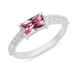 Puede incluir: Un anillo de plata con una piedra preciosa rosa de corte rectangular tipo baguette engastada horizontalmente en el centro. La banda está hecha de plata pulida y decorada con una hilera de pequeñas piedras redondas y transparentes a los lados.