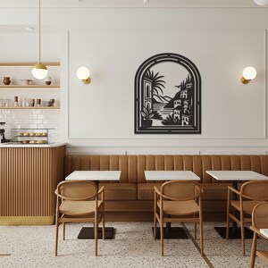 May include: A black metal wall art piece showing a coastal scene with buildings, palm trees, and a bird, framed within an arched window design. The art is mounted on a white wall in a cafe setting, above a brown leather bench and tables.