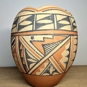 May include: A hand-painted ceramic pot with a rounded shape and a small opening at the top. The pot features geometric designs in black, orange, and cream colors. The base is a darker terracotta color.