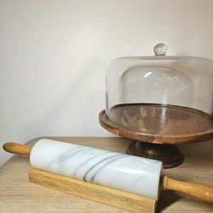 May include: A white marble rolling pin with wooden handles rests on a wooden stand. A glass cake dome sits on a wooden pedestal in the background. The rolling pin is a kitchen tool.
