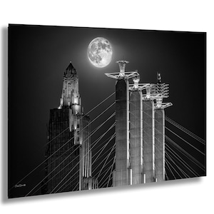 Op de afbeelding: Zwart-witfoto van een stadsgezicht 's nachts, met een volle maan. De afbeelding toont een hoog, sierlijk gebouw en een brug met verlichte torens en hangkabels. De lucht is donker en de maan is helder.