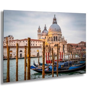 May include: A scenic photograph of Venice, Italy, featuring the Basilica di Santa Maria della Salute. The image showcases gondolas docked near wooden poles, with the church's domes and architecture in the background. The water reflects the soft light of the sky.