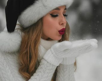 Könnte beinhalten: Eine Person trägt einen schwarzen Weihnachtsmannhut mit weißem Fellbesatz, einen weißen Pullover und weiße Handschuhe. Sie pustet Schnee, mit rotem Lippenstift und langen Wimpern. Das Bild ist in einer verschneiten Winterumgebung aufgenommen.