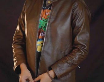 May include: A brown leather jacket with a zipper closure and a round collar. The jacket is worn over a black t-shirt with a colorful graphic design. The sleeves are long and the jacket appears to be well-fitted.