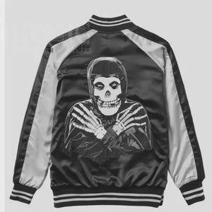 Könnte beinhalten: Schwarze Satin-Bomberjacke mit großem weißem Totenkopf-Motiv auf dem Rücken. Die Jacke hat weiß-schwarz gestreifte Bündchen und Kragen sowie weiße Einsätze an den Ärmeln. Ein klassischer, kantiger Stil.